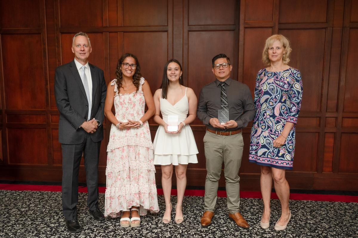 A group of five people gather wearing dresses or business suits. The three in the center hold glass awards.