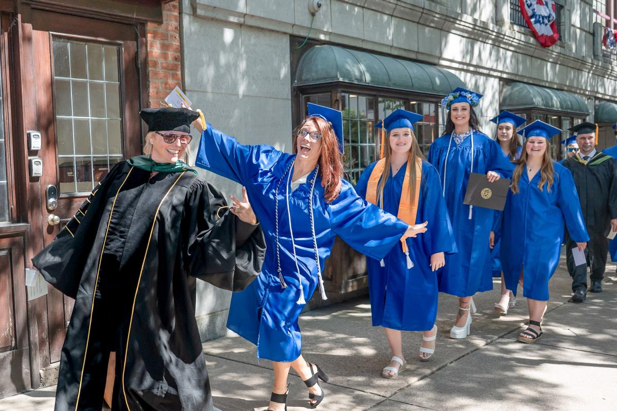 Women in graduation caps and gowns make celebratory gestures toward the camera while walking in a line down the street.