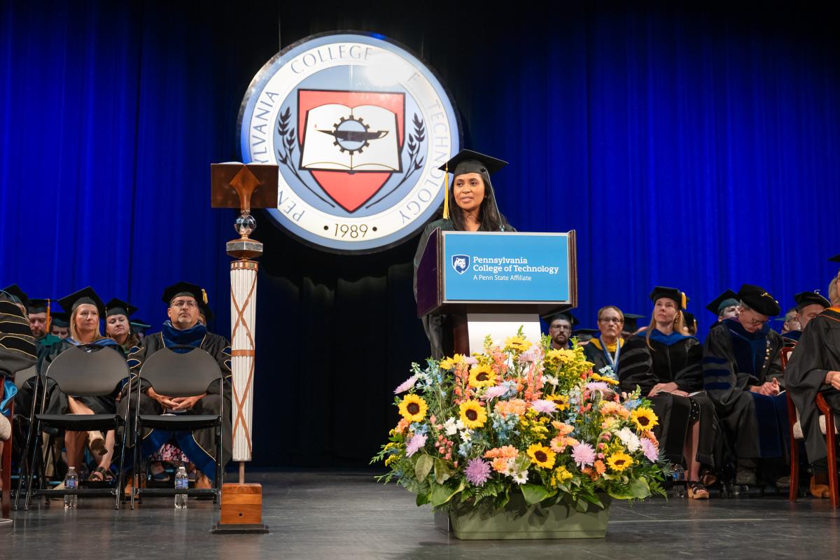 A woman in a black graduation cap and gown stands behind a podium on the Journey Bank Community Arts Center stage. Sitting behind her are many faculty members wearing caps and gowns. The Pennsylvania College of Technology seal hangs in the back, against a blue curtain.