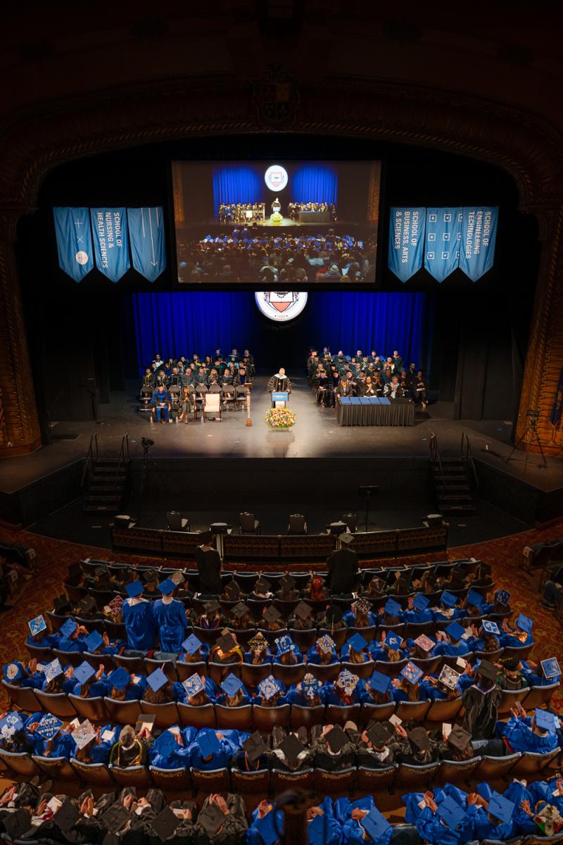 Looking down on an auditorium filled with people. On stage, a person stands behind a podium and several Pennsylvania College of Technology faculty members sit in rows in chairs behind the speaker.