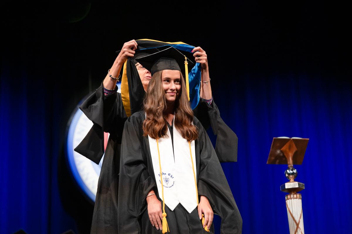 A woman in a graduation cap and gown kneals as a person behind her reaches to place an academic hood over her head.