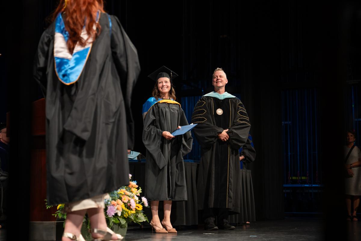 A graduate walks across the Journey Bank Community Arts Center stage toward Pennsylvania College of Technology physician assistant Jennifer Chestnut and President Michael J. Reed.