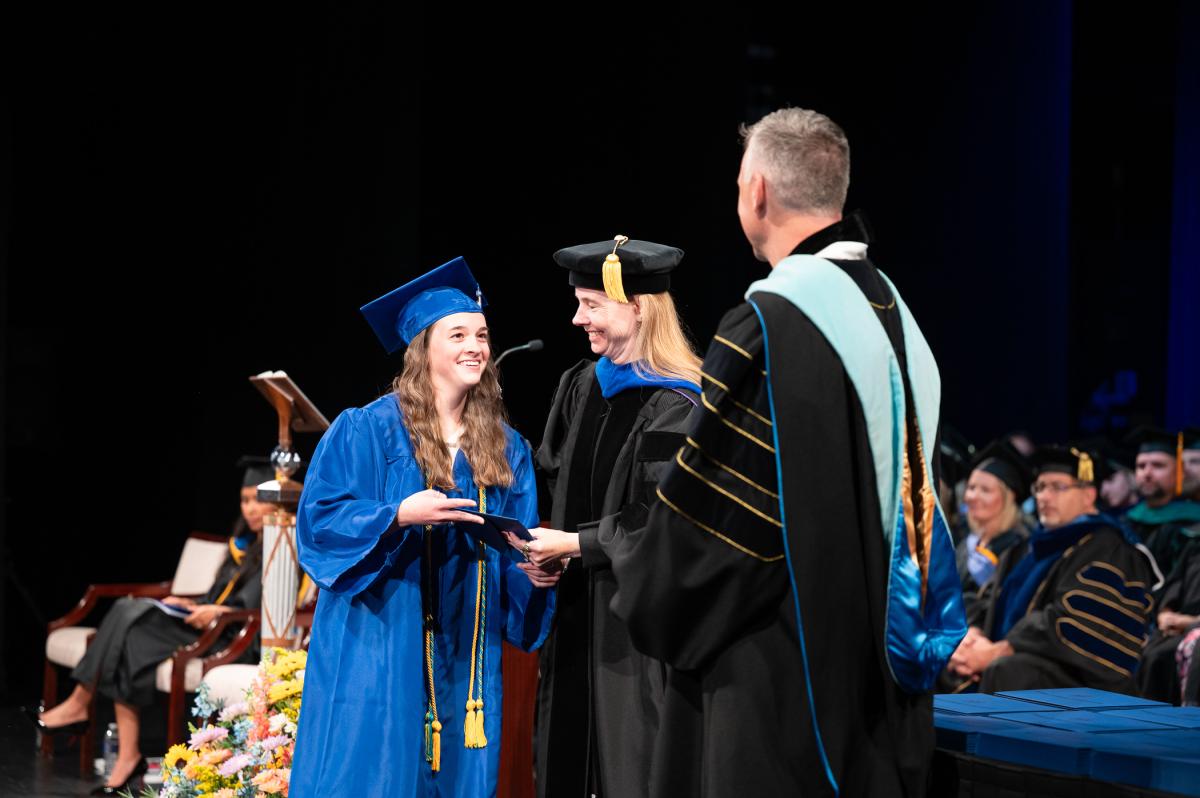 A graduate wearing a blue graduation cap and gown shakes the hand of two others in caps and gowns.