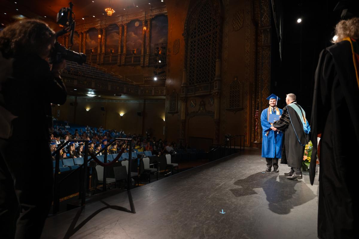 A photographer stands behind a camera while a graduate shakes the hand of Penn College President Michael J. Reed.