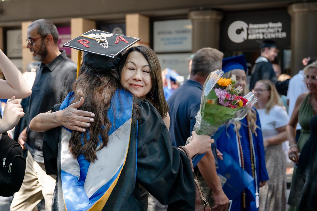 A graduate wearing a graduate cap and gown hugs a woman outside the Journey Bank Community Arts Center.