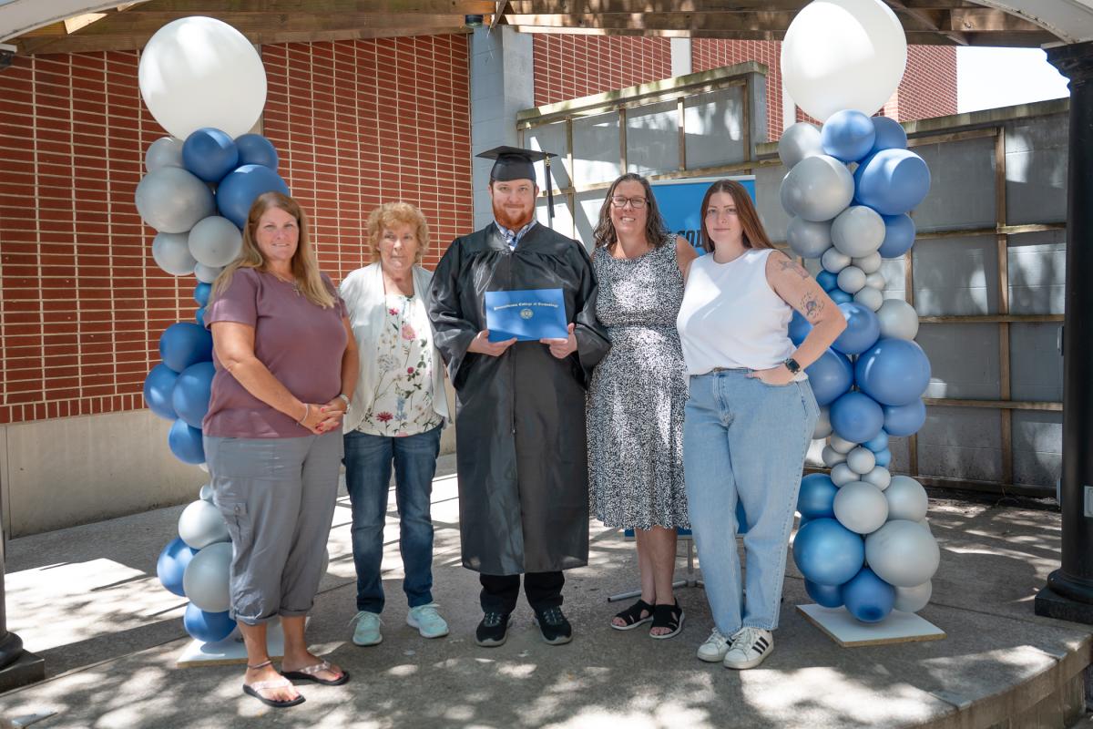 A man in a black graduation cap and gown holds a blue diploma folder. He is flanked by four family members and two pillars of blue and gray balloons.
