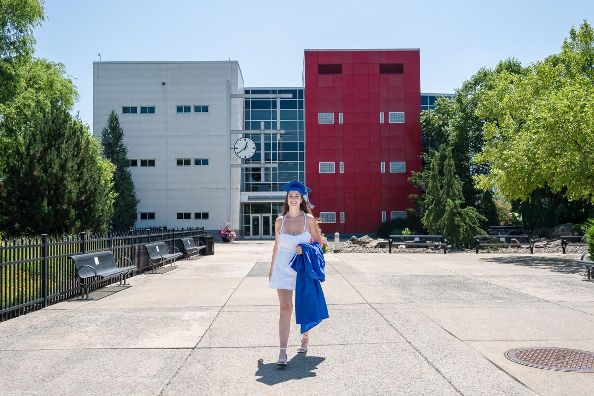 A woman in a white dress holds a blue graduation gown and stands in front of the Pennsylvania College of Technology Madigan Library.