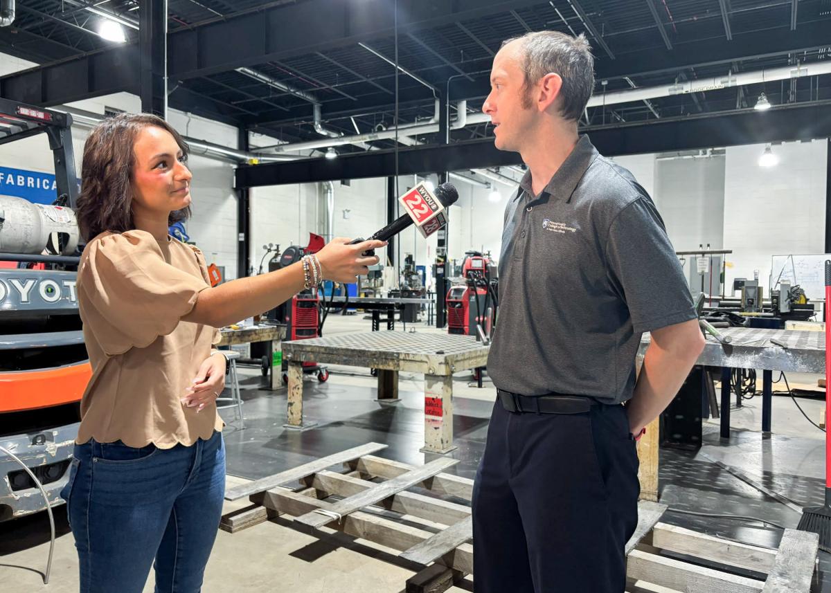 A woman holds a microphone up to a man inside Pennsylvania College of Technology's Lycoming Engines Metal Trades Center. Industrial equipment is in the background.