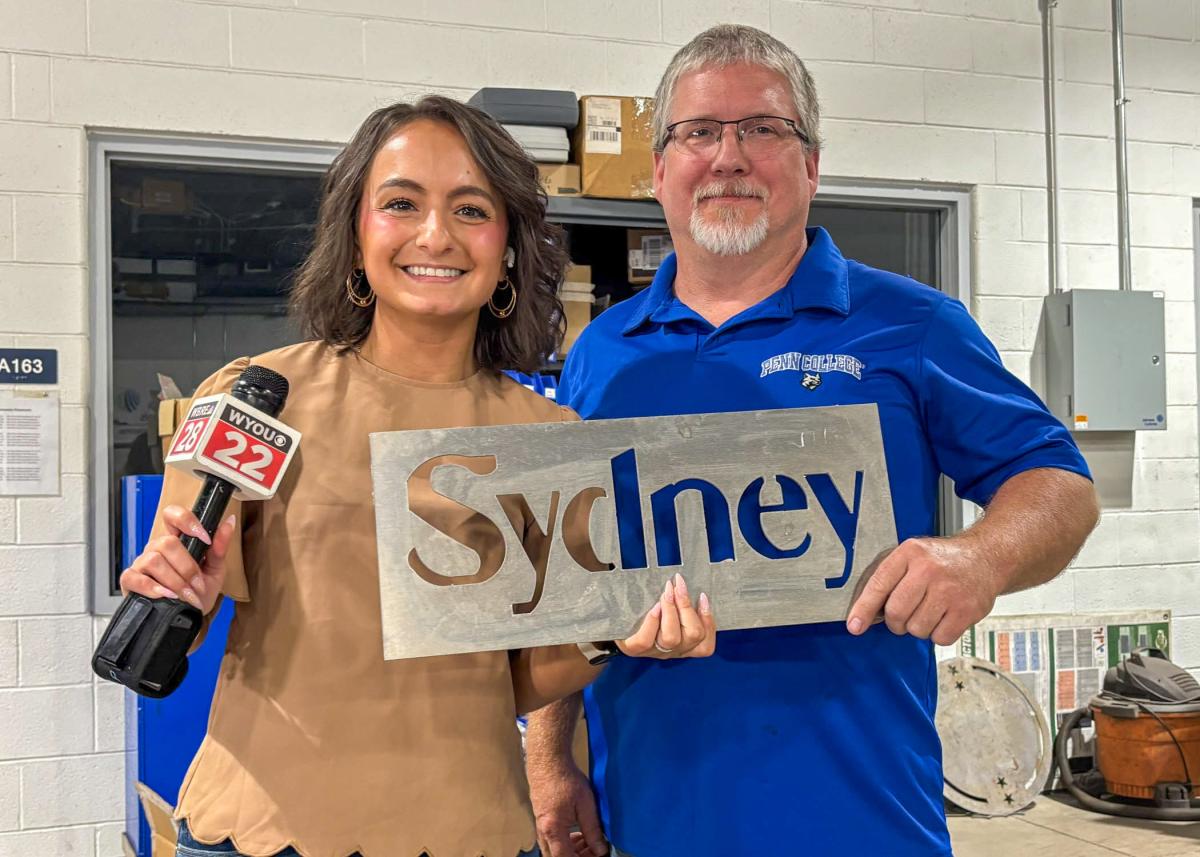 A woman with a microphone stands next to a man. Both are holding a metal sign that says "Sydney" in cut-out letters.