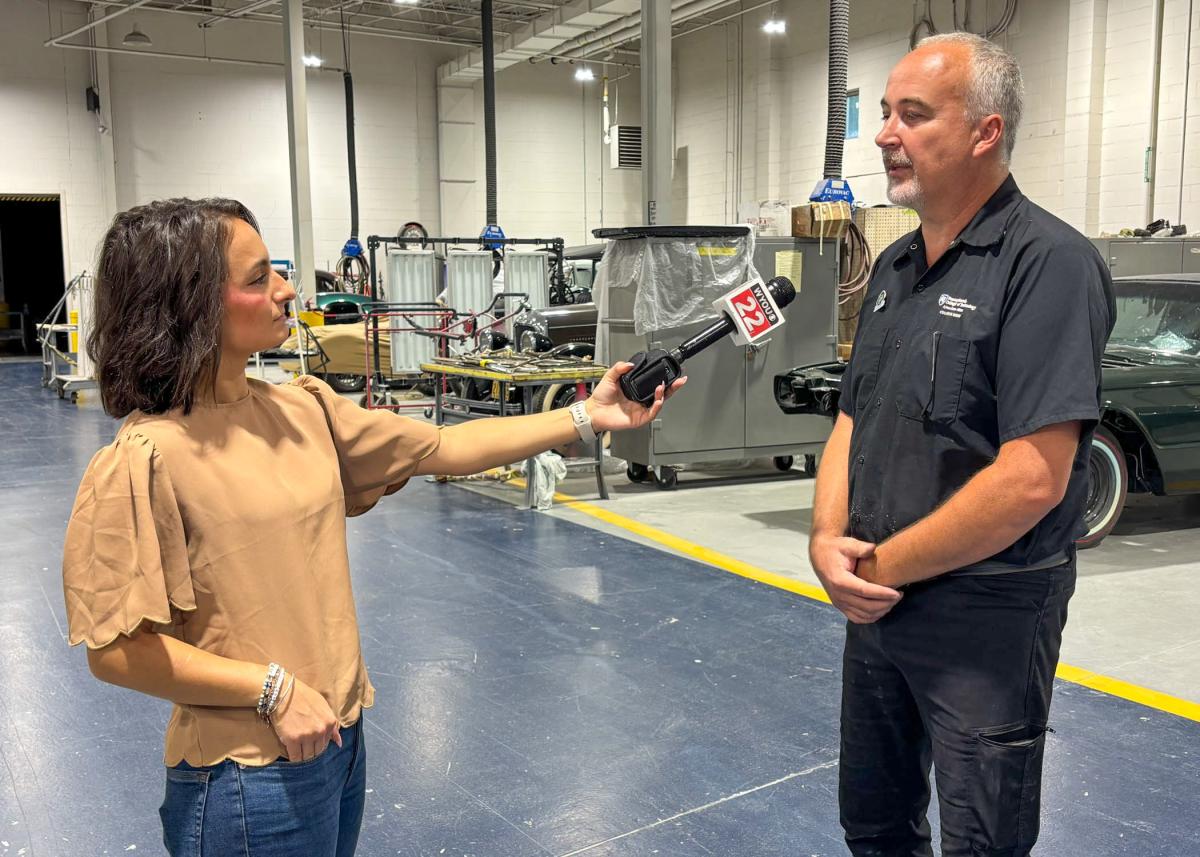A woman holds a microphone up to a man inside Pennsylvania College of Technology's collision repair lab. Antique cars are in the background.