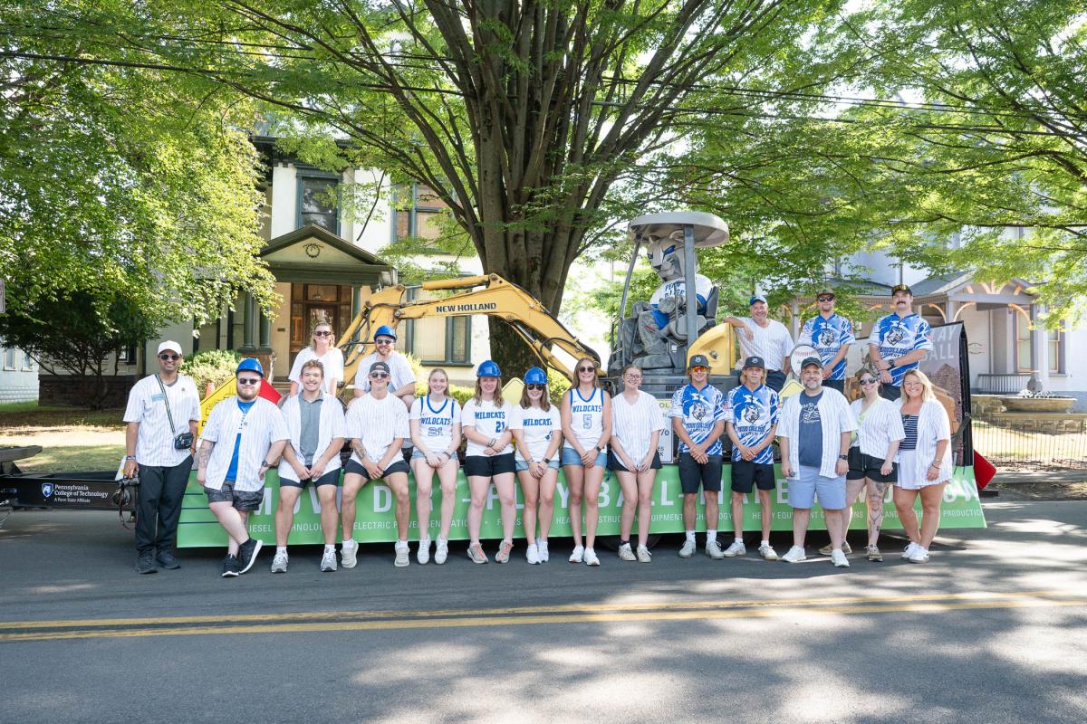 A large group of people stands in front of a trailer that is decorated with signs and is carrying an excavator. The Penn College Wildcat mascot is sitting in the excavator.