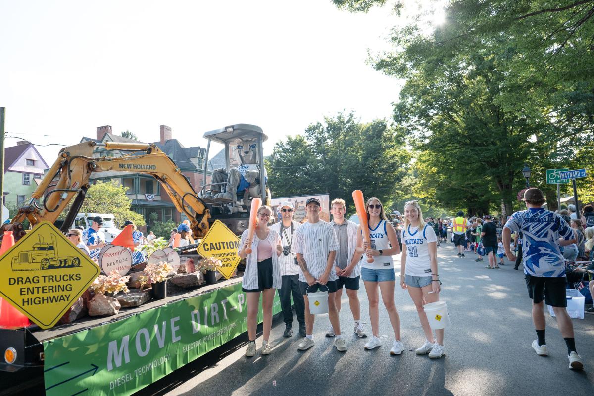 A group of six Penn College representatives wearing sports uniforms stands next to the Penn College float, with the Wildcat mascot sitting in an excavator.