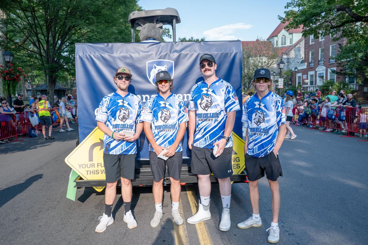 Four young men in shirts that say Penn College Archery stand behind the college's float, on a street lined with people.