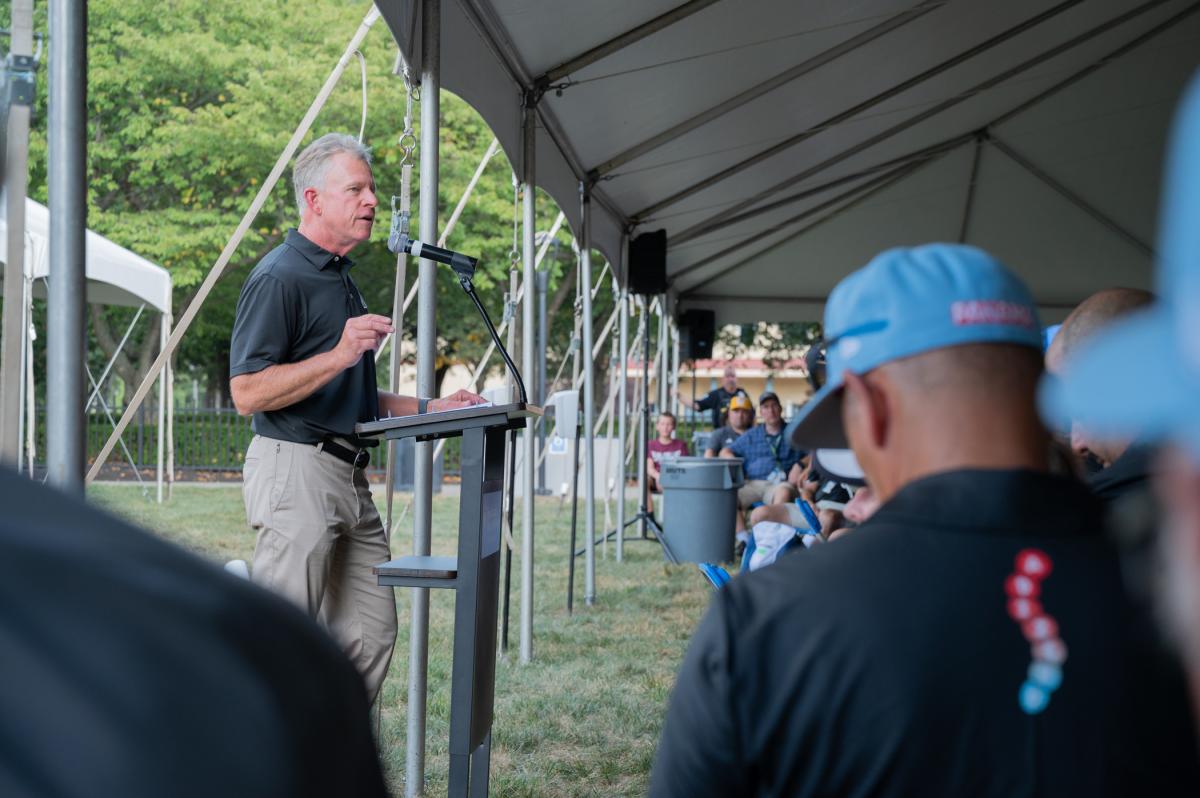 A man in Vanderbilt University polo shirt talks into a microphone as people in baseball uniforms listen.