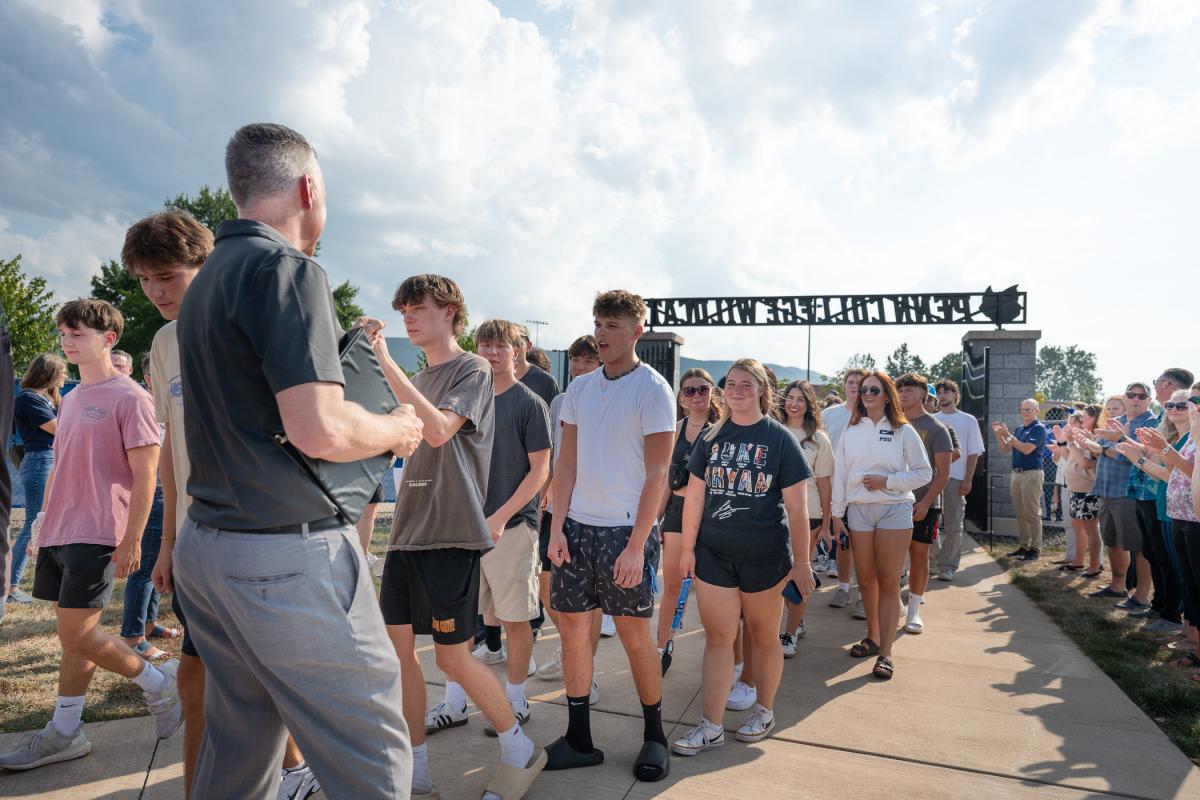 A line of students enters UPMC Field, cheered by employees on the sideline. In the foreground, the college president gives fist bumps to those walking past.