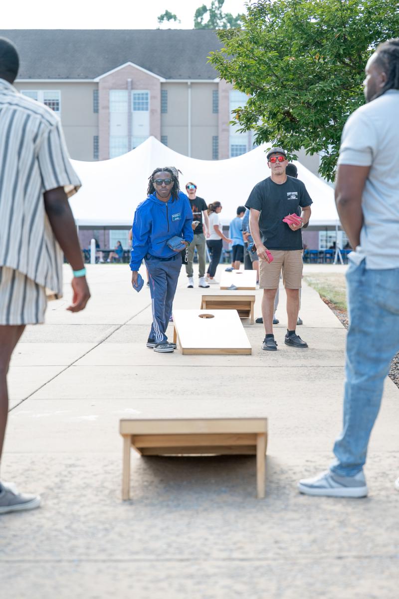 A student prepares to throw a bean bag at a cornhole board, while three others watch.