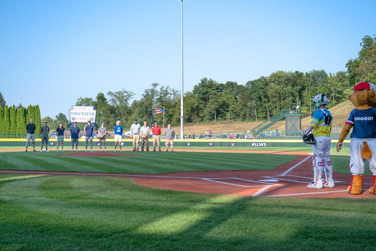A group of Penn College representatives stands at mid-field at Volunteer Stadium, while a young catcher and Dugout, Little League's mascot, stand near home plate.