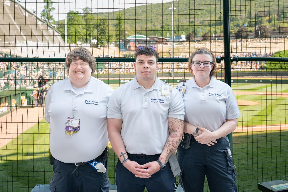 Three people in paramedic uniforms stand in front of a fense. Behind the fence is a baseball game.