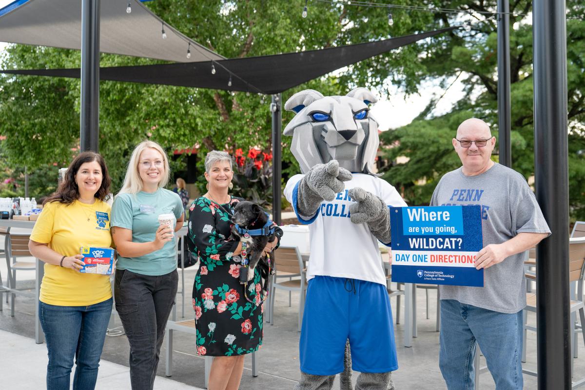 Four people and the Wildcat mascot look at the camera. One is holding a box of Pop-Tarts, one is holding a disposable coffee cup, one his holding a dog, and one is holding a sign that reads: Where are you going, Wildcat? Day One directions."