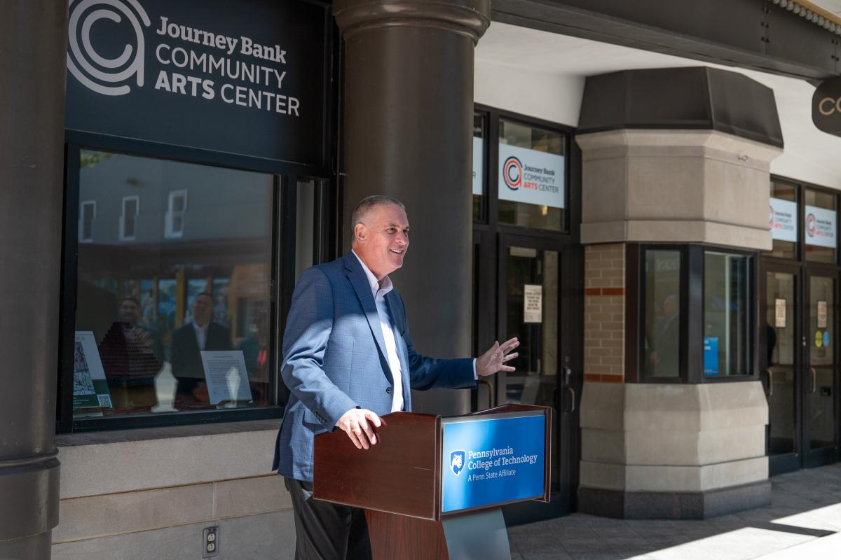 A man in a business suit stands behind a podium outside the Journey Bank Community Arts Center.