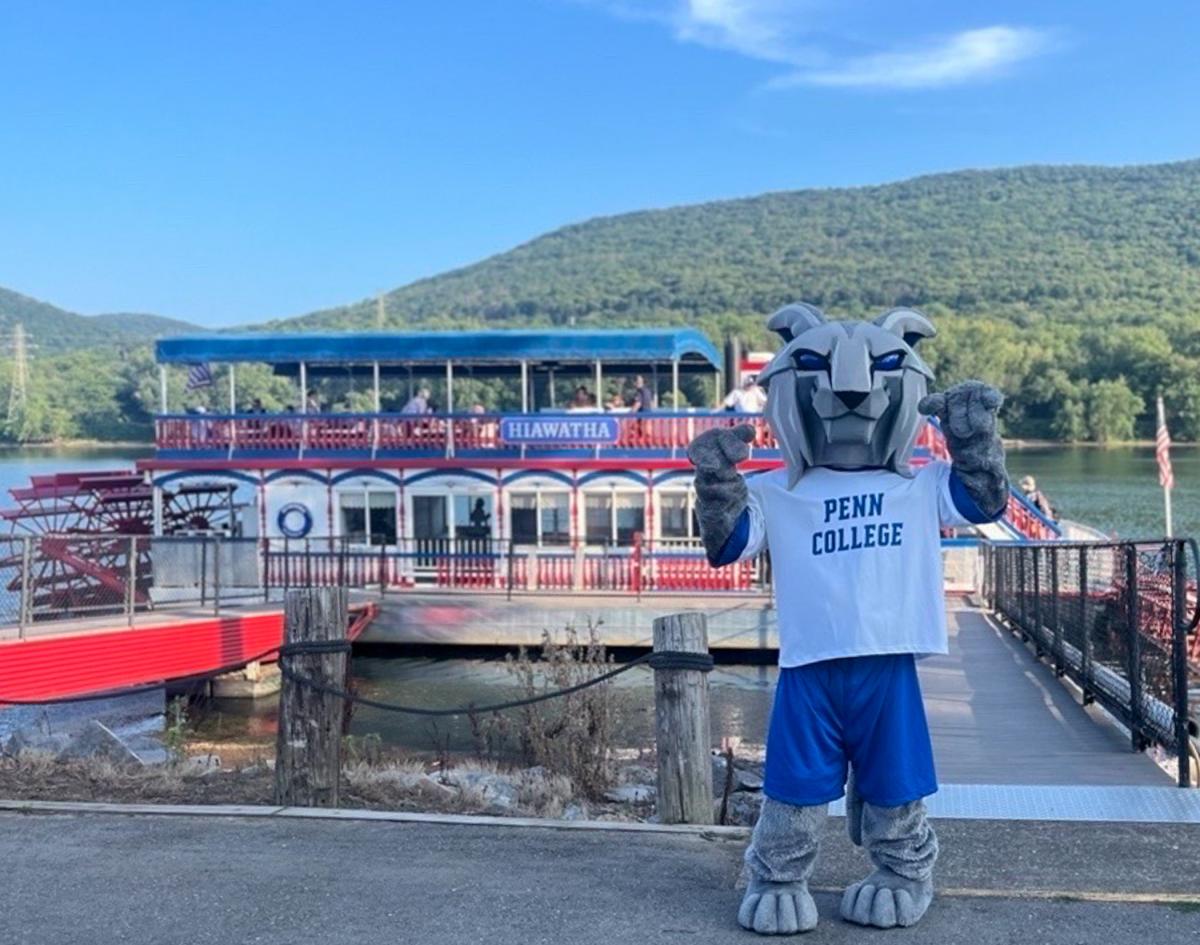Penn College's mascot, the Wildcat, stands in front of the Hiawatha Paddlewheel Riverboat docks. The white riverboat with red trim and red paddlewheel are behind the Wildcat.
