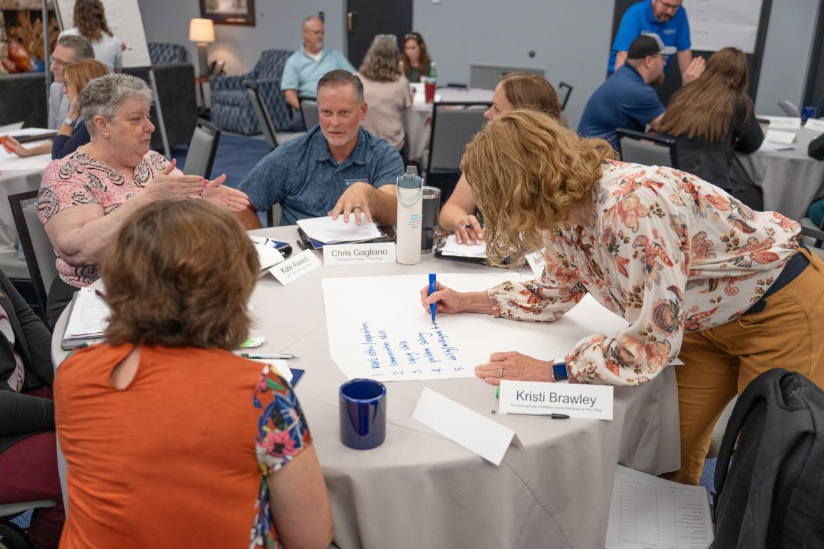 People in business attire sit around a table and engage in conversation while one writes on a large sheet of paper.