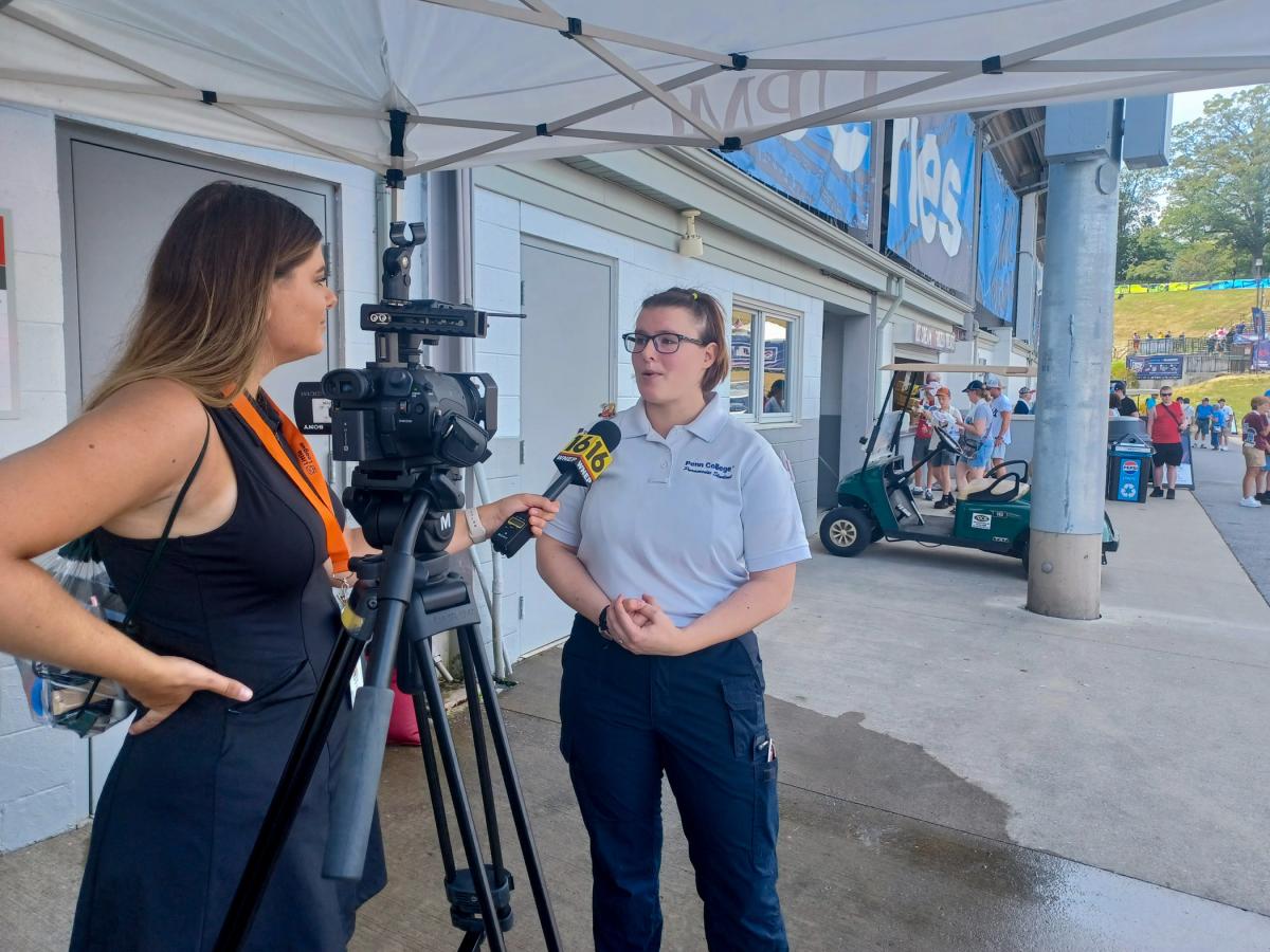 A woman holds up a microphone to another woman in paramedic uniform. Between them is a television camera. Behind them are the walls of Lamade Stadium at the Little League International Complex.
