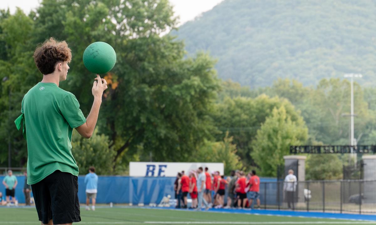 A student in green spins a kickball on his finger while a group in red shirts gathers in the far background.