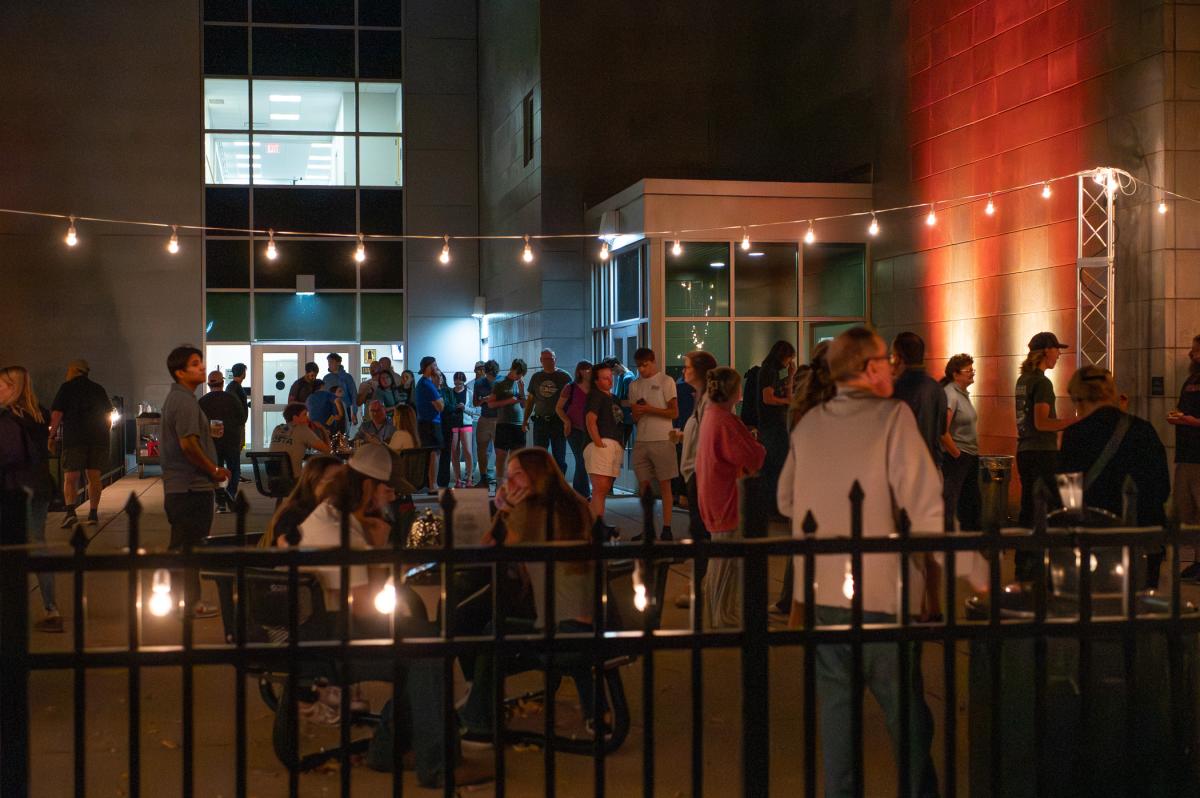 A large group of people stands and sits at tables outside Penn College's Madigan Library, where string lights are hung.