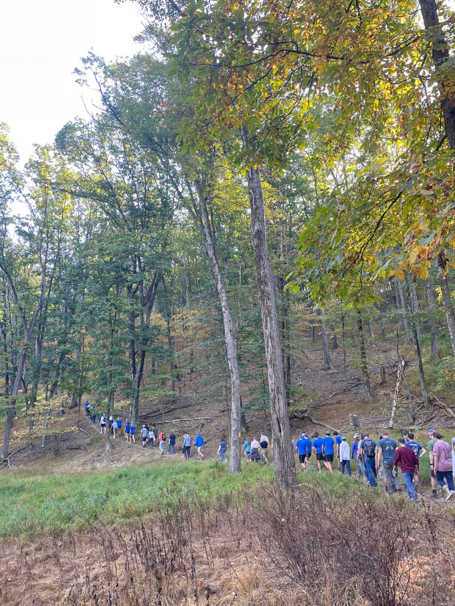 A long line of people walks up a narrow wooded trail.