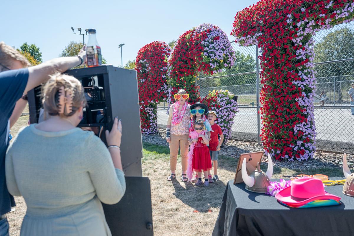 Four children wear bright, oversized sunglasses and other costume props while standing in front of giant live-flower sign that spells out PCT.  Adults look through a camera at them.