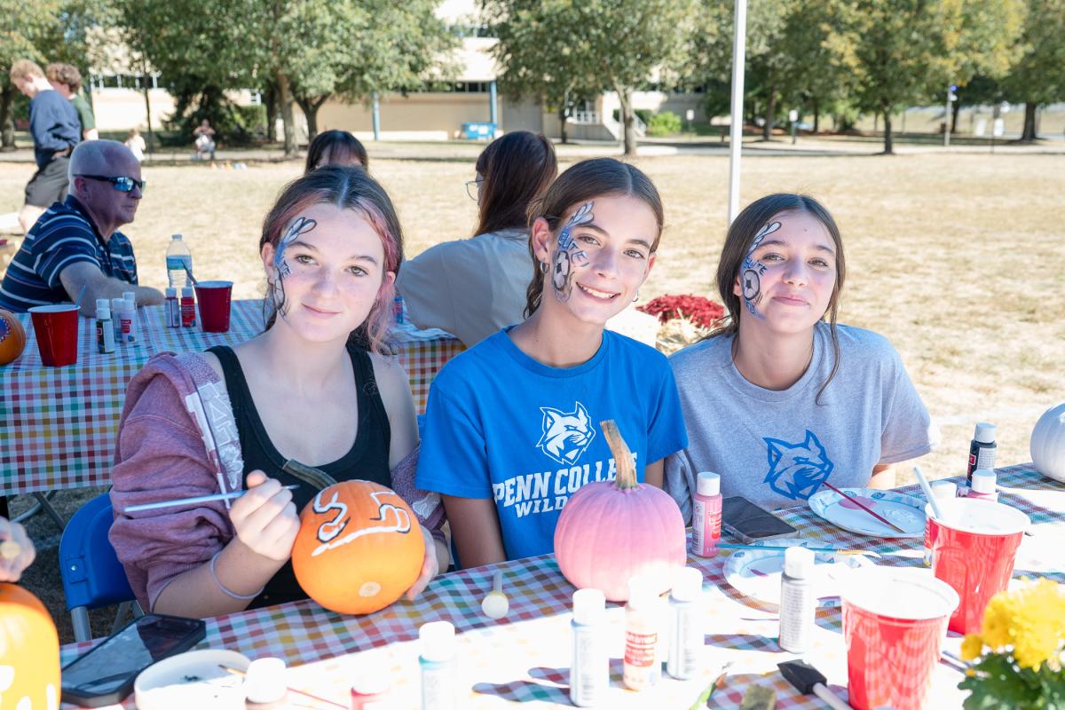 Three young women with matching soccer-themed designs painted on their cheeks look at the camera. They are sitting at a table with paint brushes, paint bottles and painted pumpkins. One is holding her paintbrush and partly painted pumpkin.