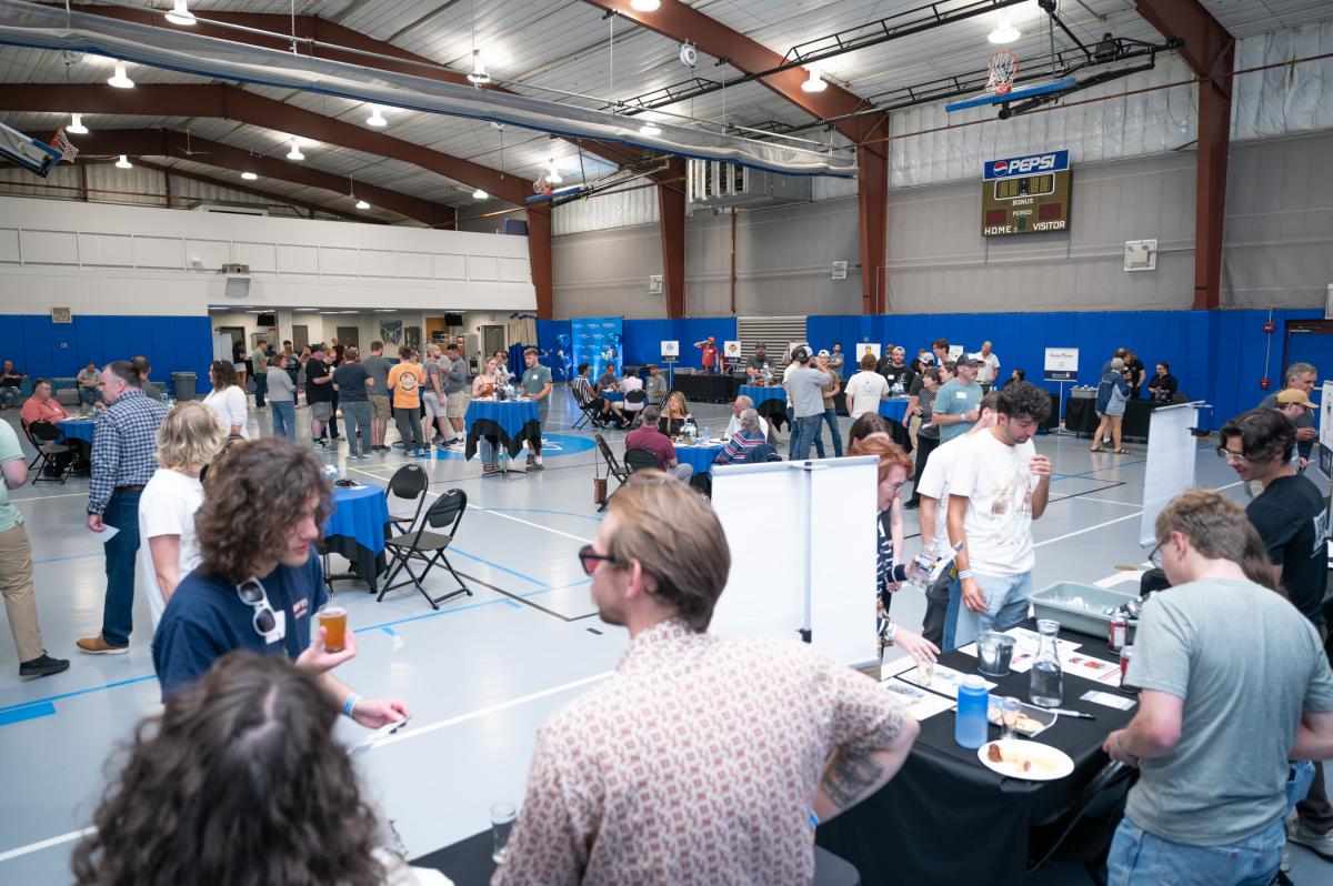 In teh foreground, a line of tables offer beer samples. In the background, groups of people gather at tables.