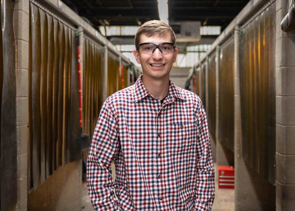 Clayton Fegley stands among welding booths. He's wearing safety glasses.