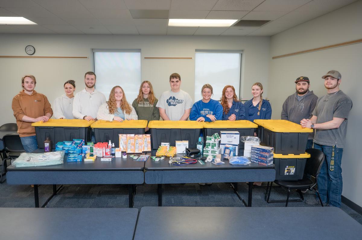 A group of students stands behind a table filled with large plastic storage totes and medical supplies.