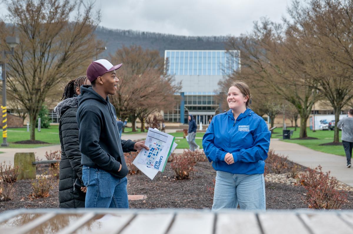 A prospective student, holding campus maps, speaks to a smiling Morgan Bartholomew on the campus mall, with the Advanced Technology & Health Sciences Center in the background.