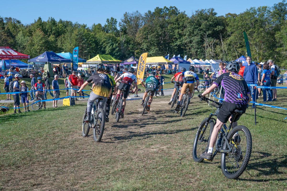Bicyclists round a corner. Colorful team and sponsor tents are in the background.