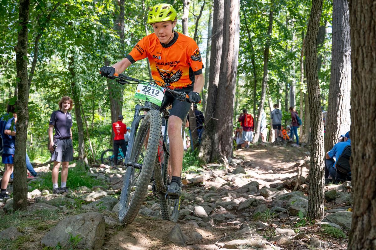 A boy rides a bike down a rocky, wooded path while onlookers stand along the edges of the trail.