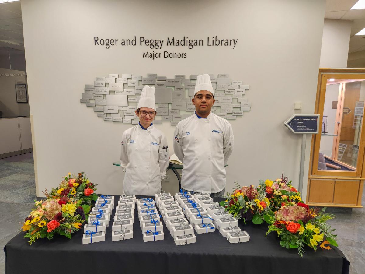 Two students in chefs uniforms stand behind a table. Rows of small white boxes are on the table.