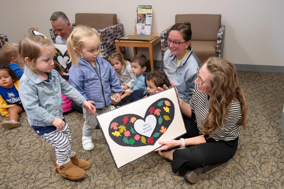 Two young children hand an oversized handmade card to a woman sitting on the floor while other children sit on the floor and watch.
