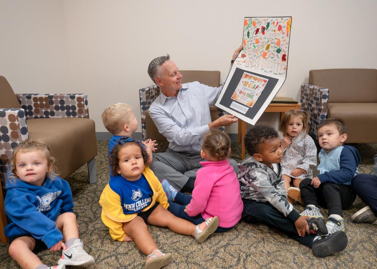 Penn College President Mike Reed sits on the floor. He is surrounded by toddlers and is looking at an oversized hand-made card.
