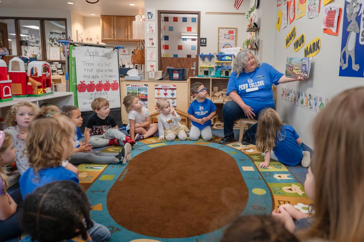 Preschool children sit in a circle on the floor around a soman who is holding a picture book.