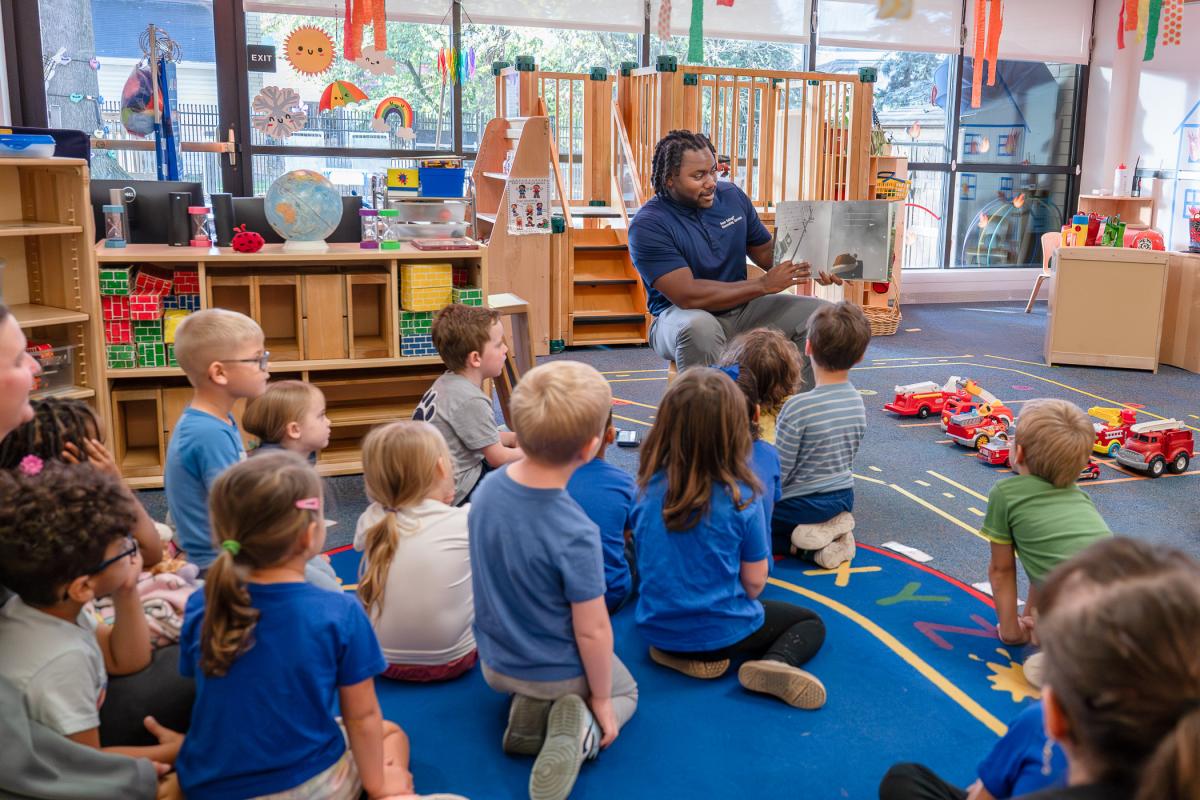 A man sits in a chair reading a picture book. Children sit on the floor looking at him and the book he is holding.