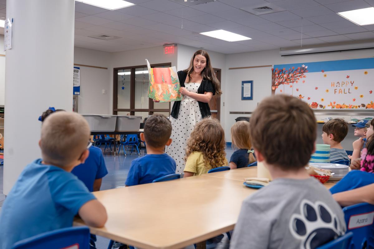 Children sit around a table in a cafeteria looking at a woman who is standing in front of them, holding and reading a large picture book.
