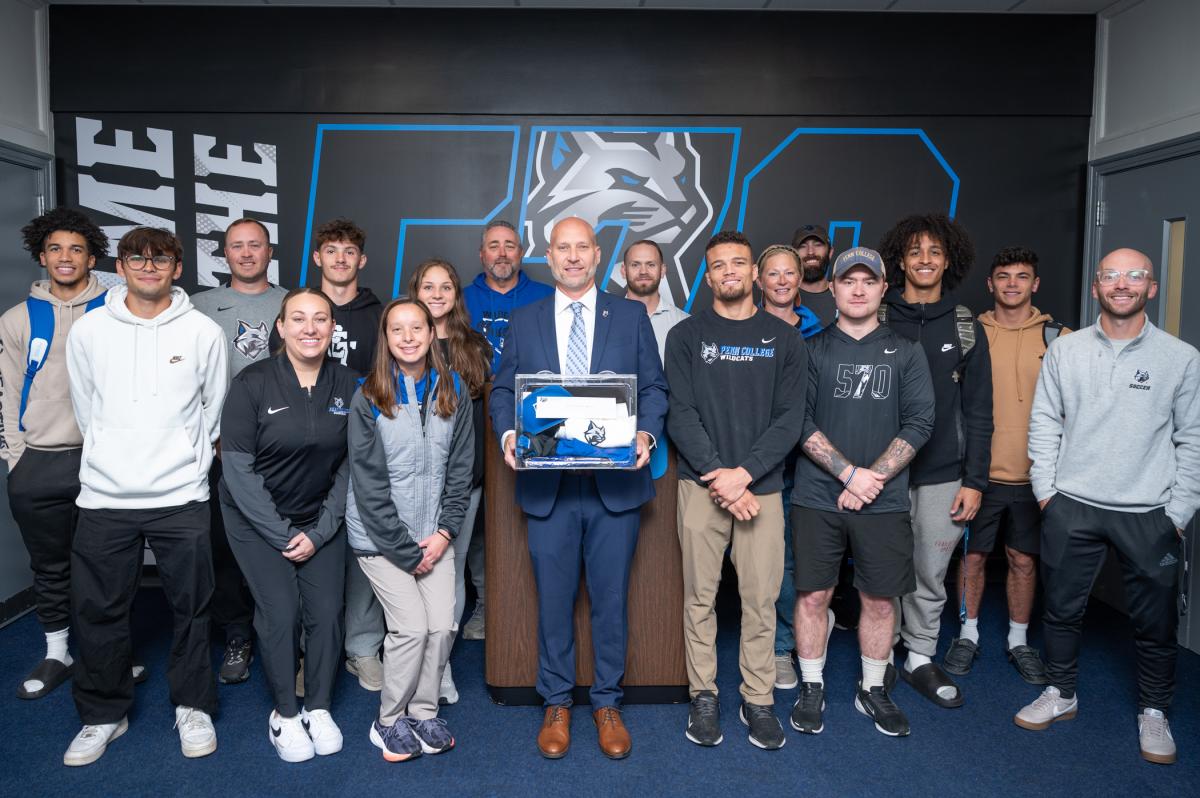 A group gathers in the Bardo Gymnasium lobby. In the center is athletic director Scott Kennell, holding a clear box filled with items.