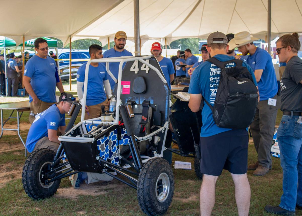 Penn College students and inspectors gather around the college's Baja car in the tented inspection area.