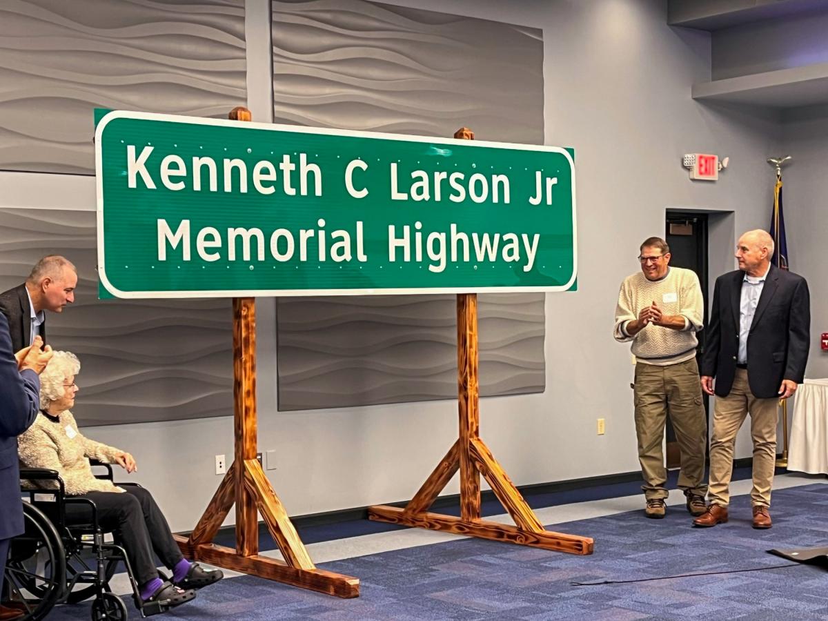 Inside Penn College's Thompson Professionald Development Center, people on either side of a highway sign applaud. The sign is hanging on wooden posts.