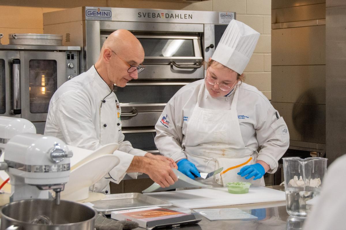 In Penn College's baking lab, a man in a chef's uniform provides instruction to a young woman, also in a chef's uniform, as both look at a product on a stainless steel table. Commercial ovens are behind them.