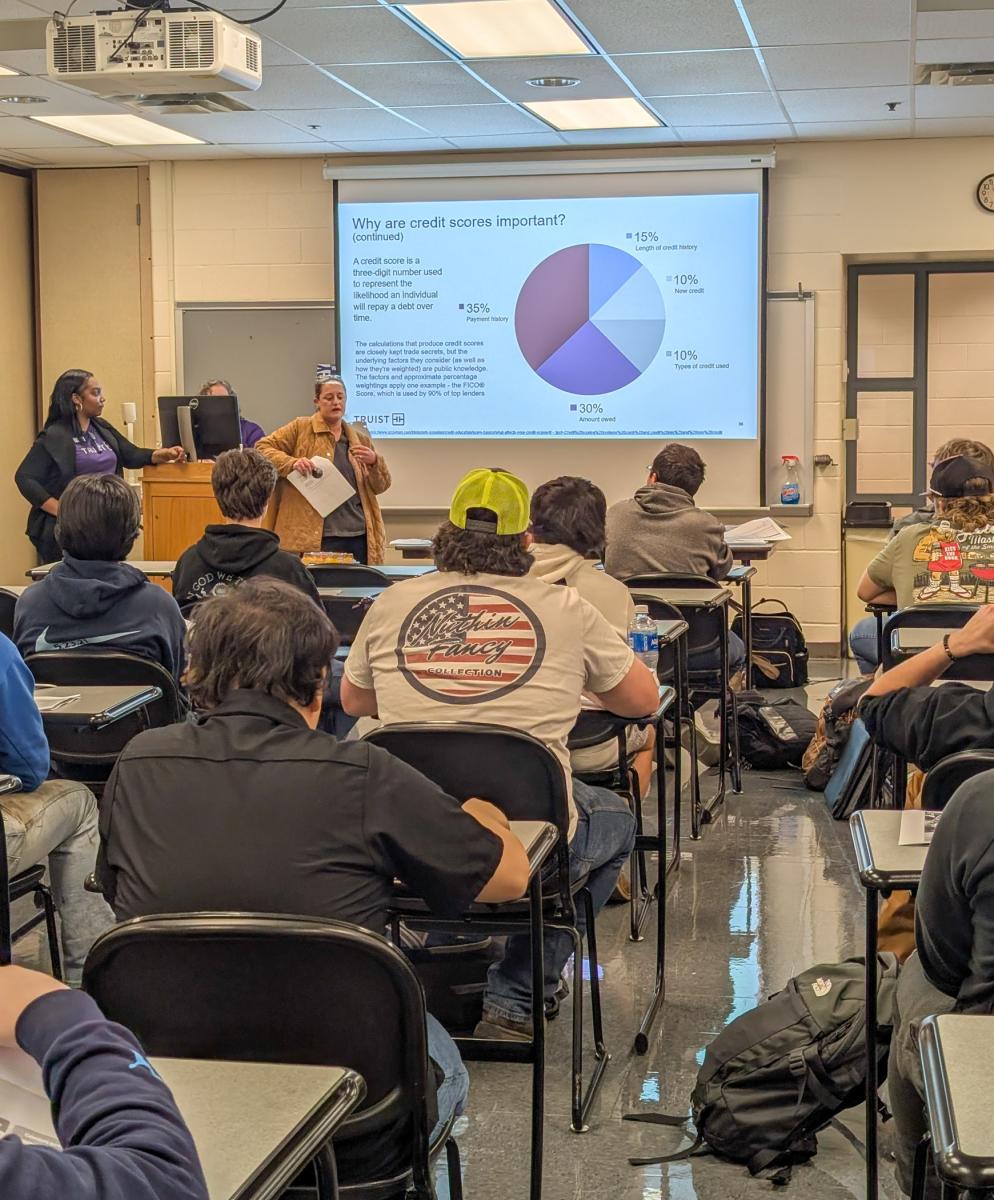 A woman speaks in front of a full classroom. Behind her is a projector screen showing a pie chart.