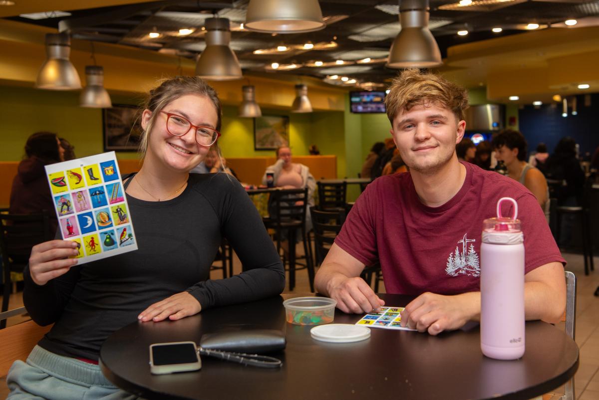 Two students sit at a table in Penn College's CC Commons. One is holding up a loteria board. The other has a board sitting in front of him. A cup of bingo markers sits between them to mark their boards.